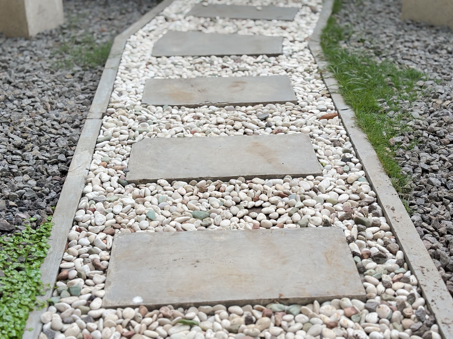 A stone walkway made of rectangular pavers set in white gravel, flanked by small patches of green grass and dark gravel.