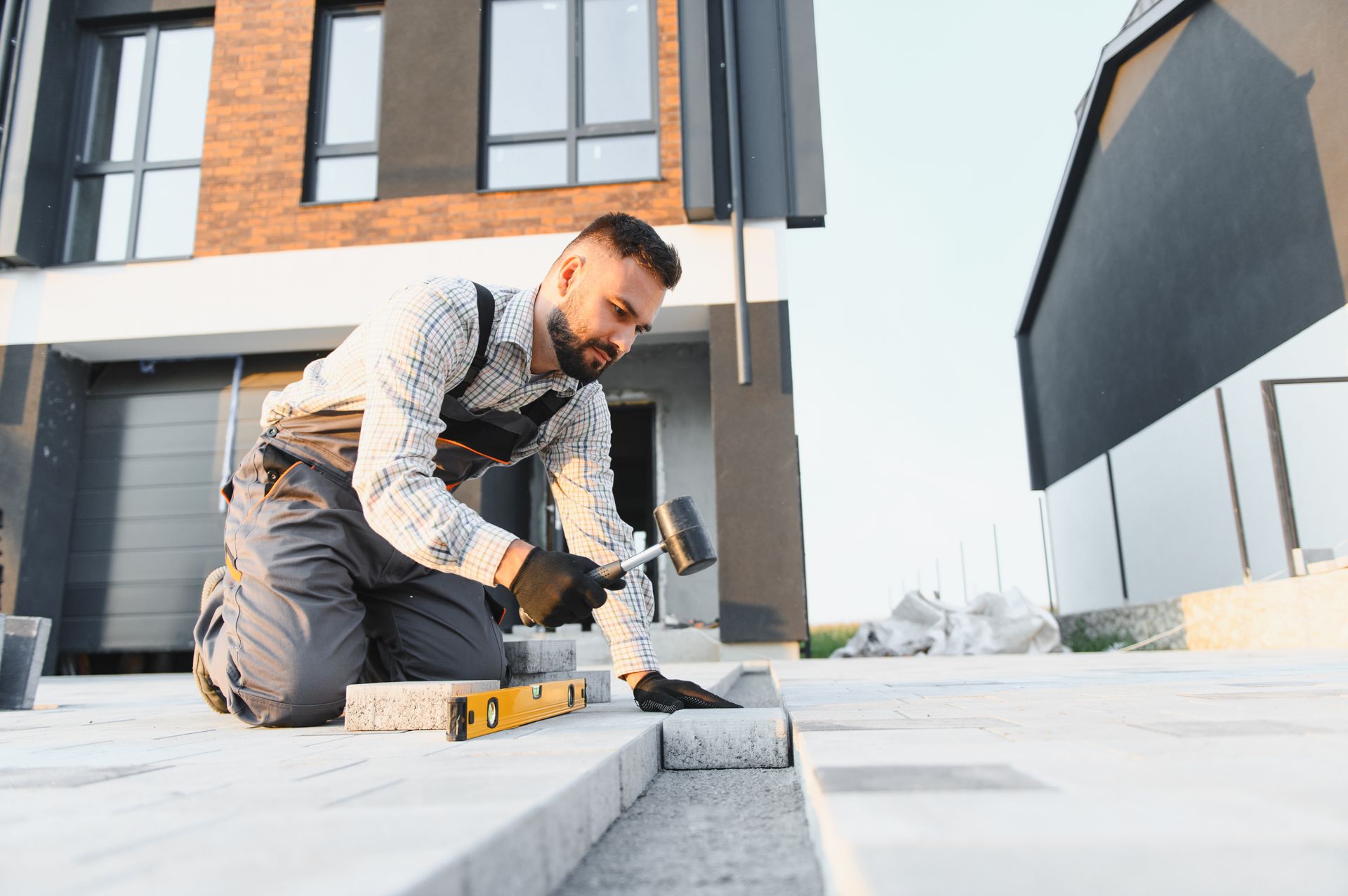 A person in work clothes kneels on a residential patio, using a mallet and level to install grey paving stones.