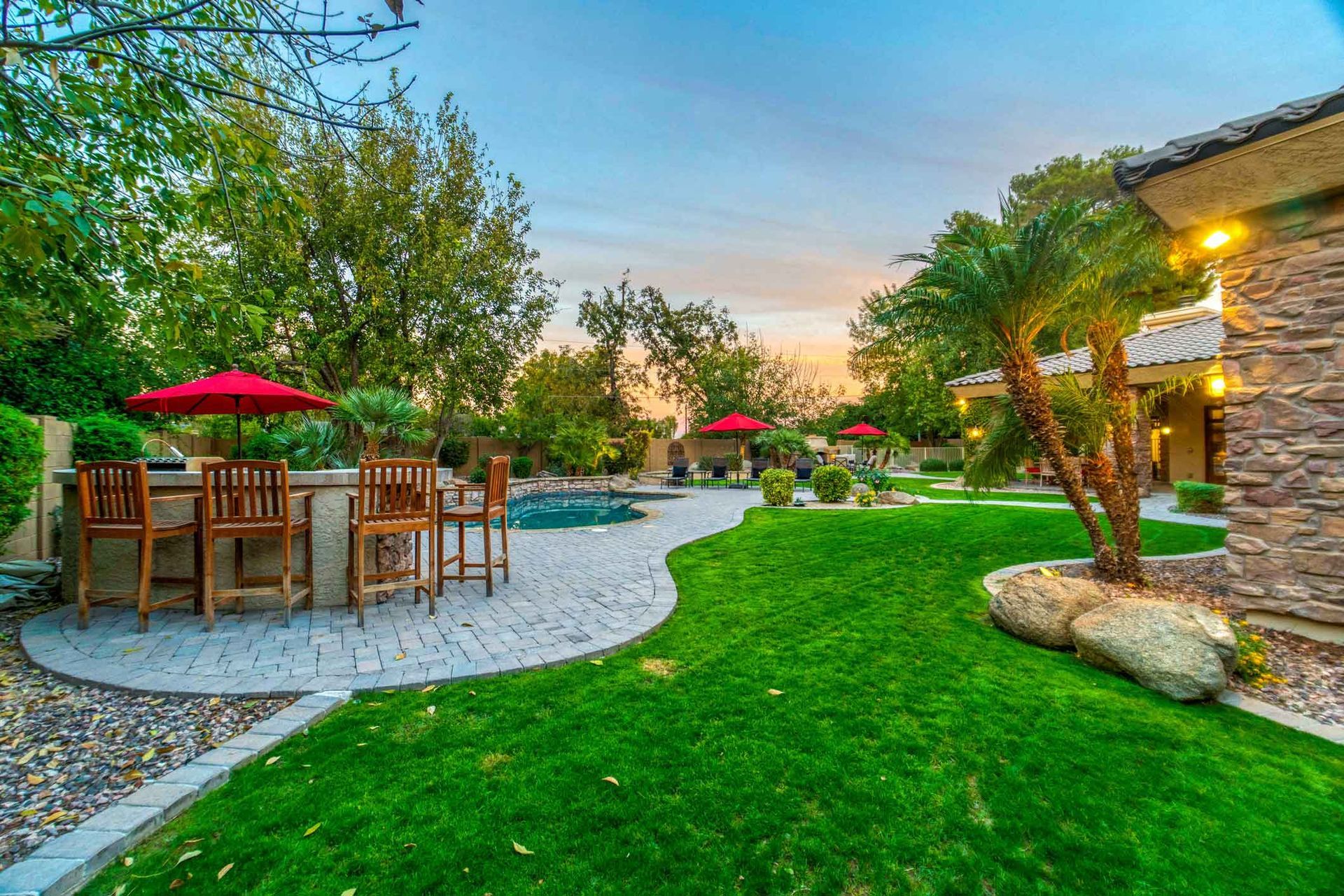 Backyard with a stone patio, bar stools, red umbrellas, green lawn, palm trees, and a view of a house at sunset.