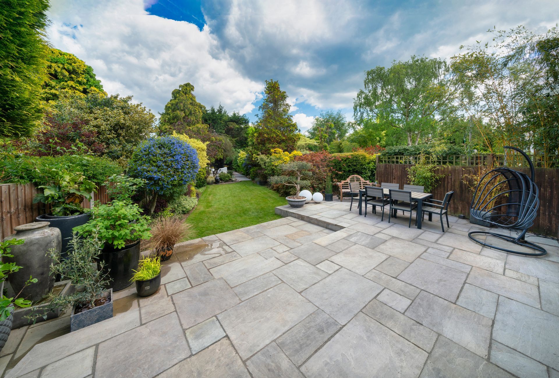 A stone patio in a lush backyard featuring outdoor dining furniture, a hanging chair, and a green lawn under cloudy skies.