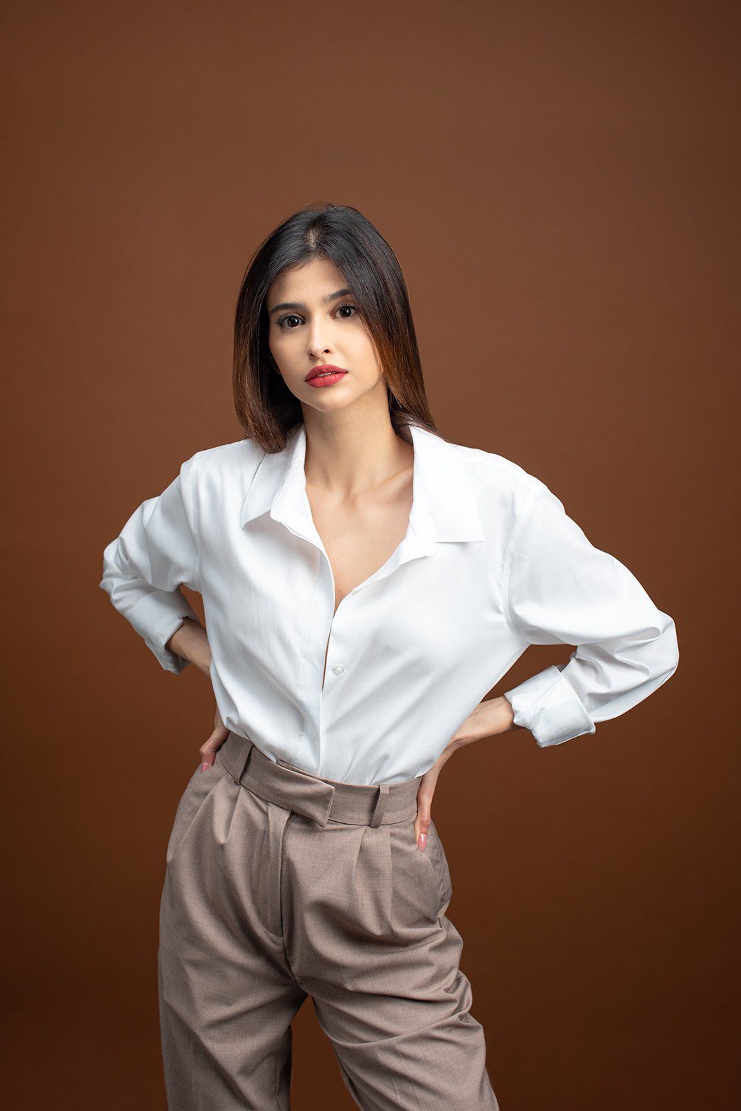 Woman in white shirt and brown pants, hands on hips, brown background.