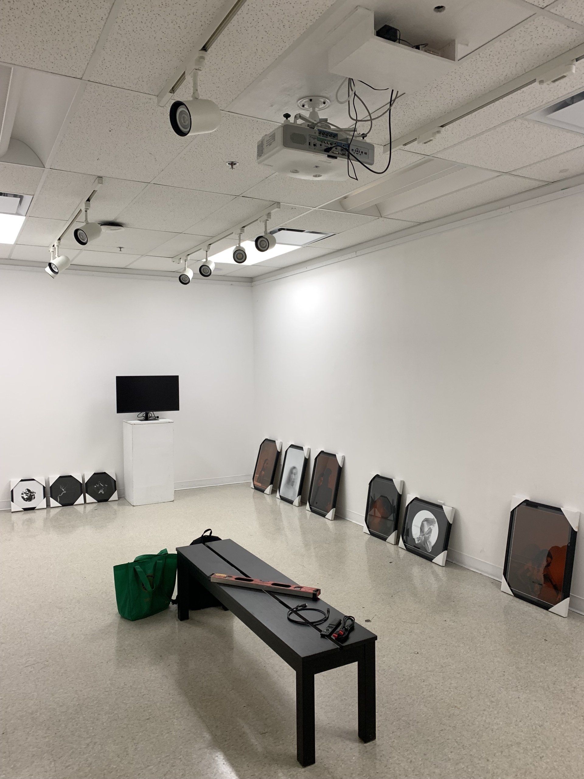 White-walled gallery with art on floor, bench, and TV screen. Projectors on ceiling.
