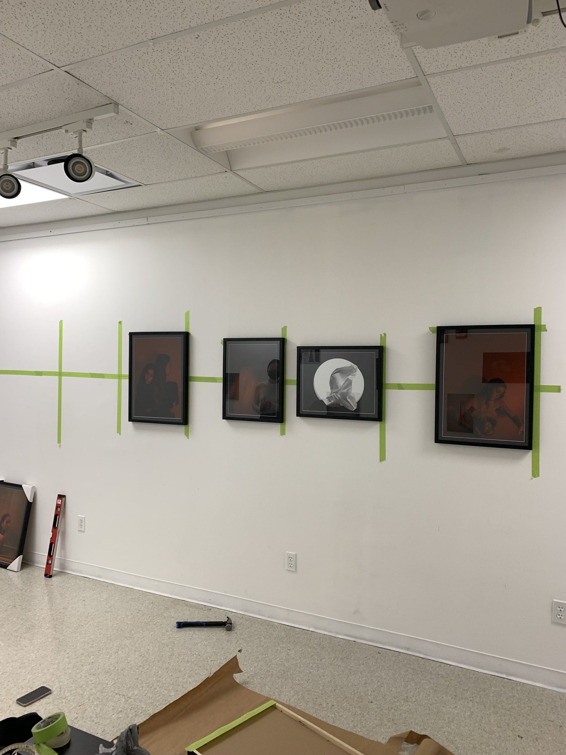 Four framed artworks taped to a white wall, level line marked with green tape. Room is empty.