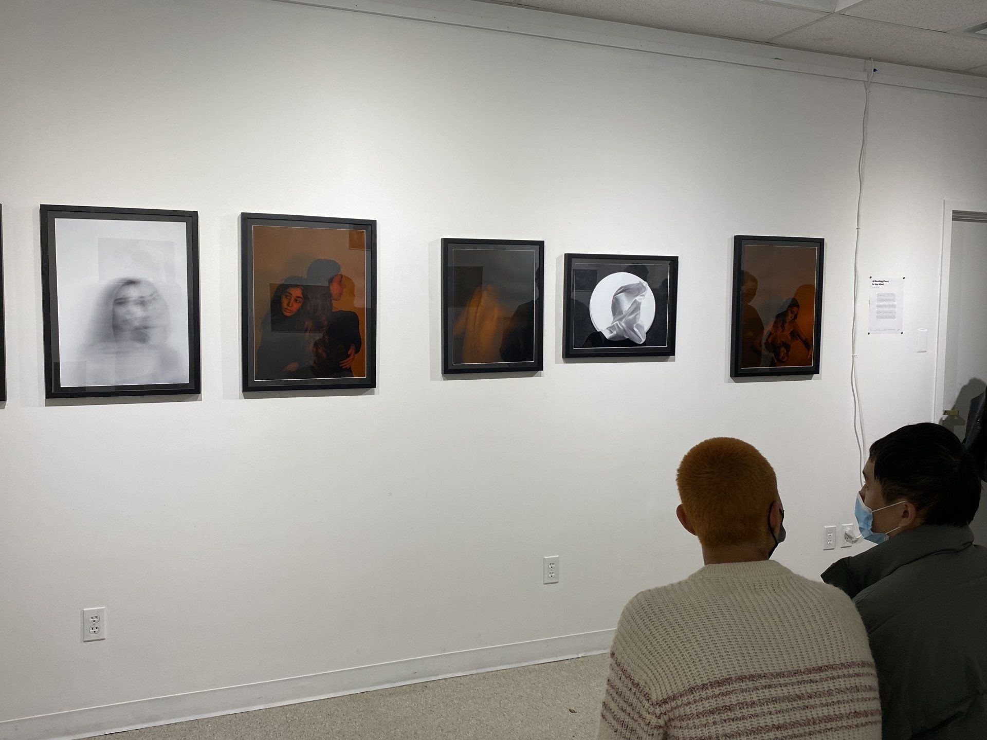 People looking at framed art on a white gallery wall. The art includes portraits with dark frames.