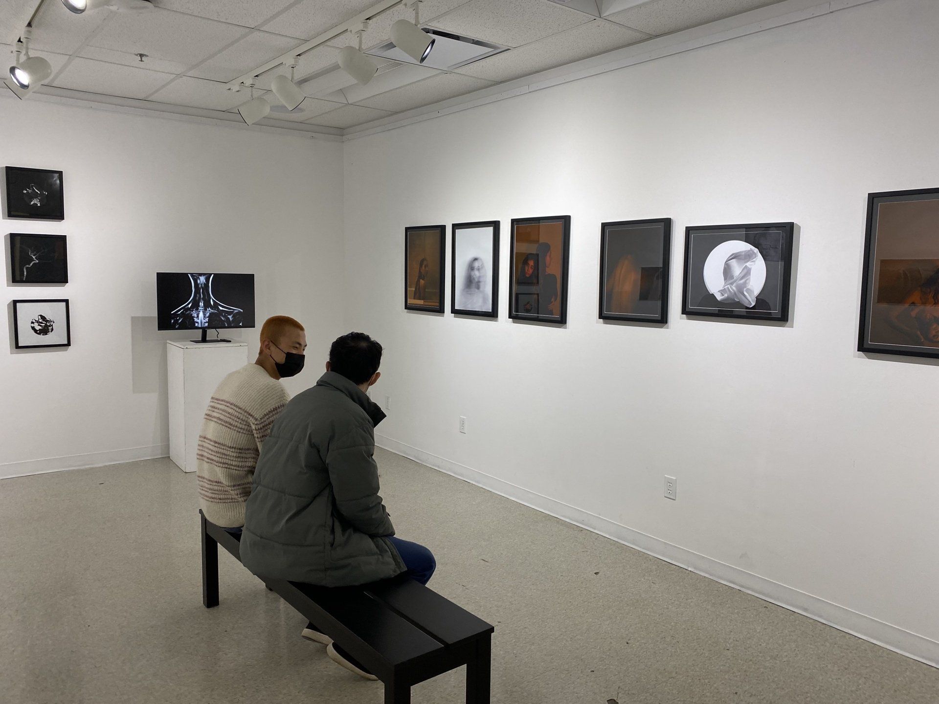 Two people sit on a bench, looking at framed artwork in a white-walled gallery. A video screen is visible.