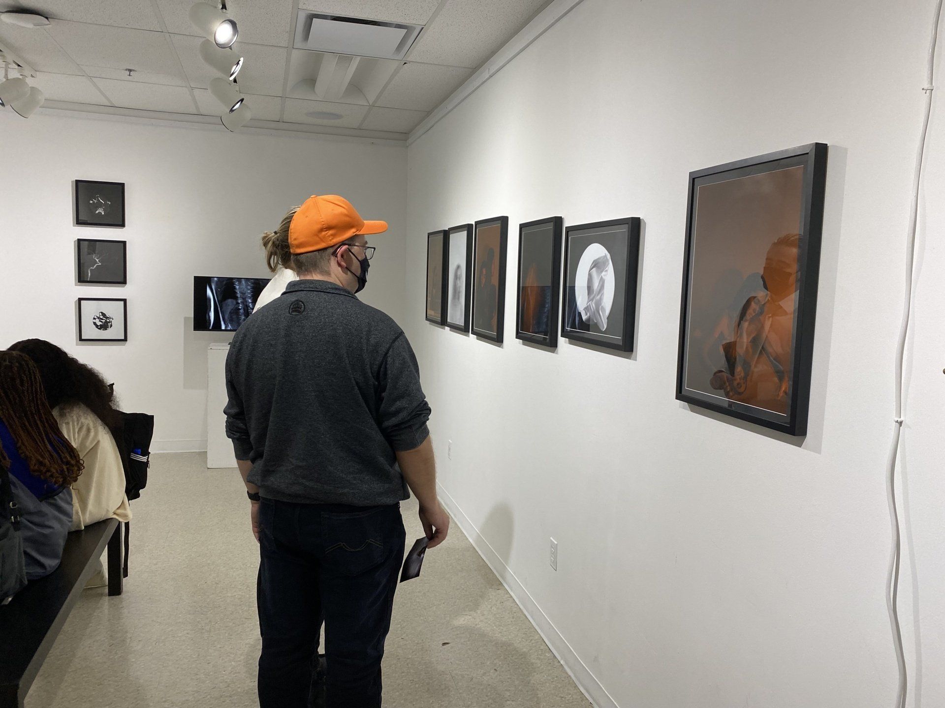 Person in an orange cap views framed artwork in a gallery. White walls, art, and a small video screen are visible.
