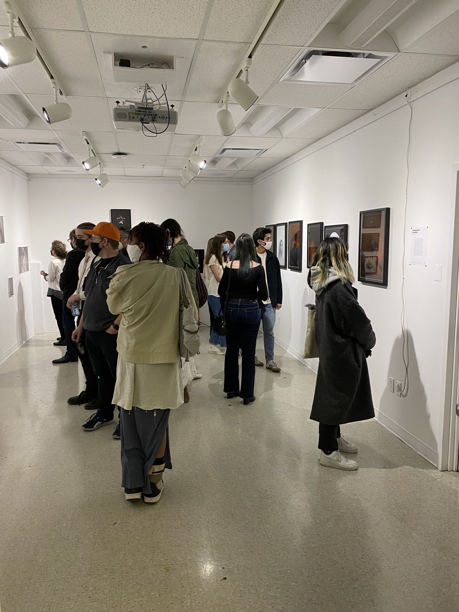 People viewing artwork in a gallery, light-colored walls, overhead lights, some wearing masks.