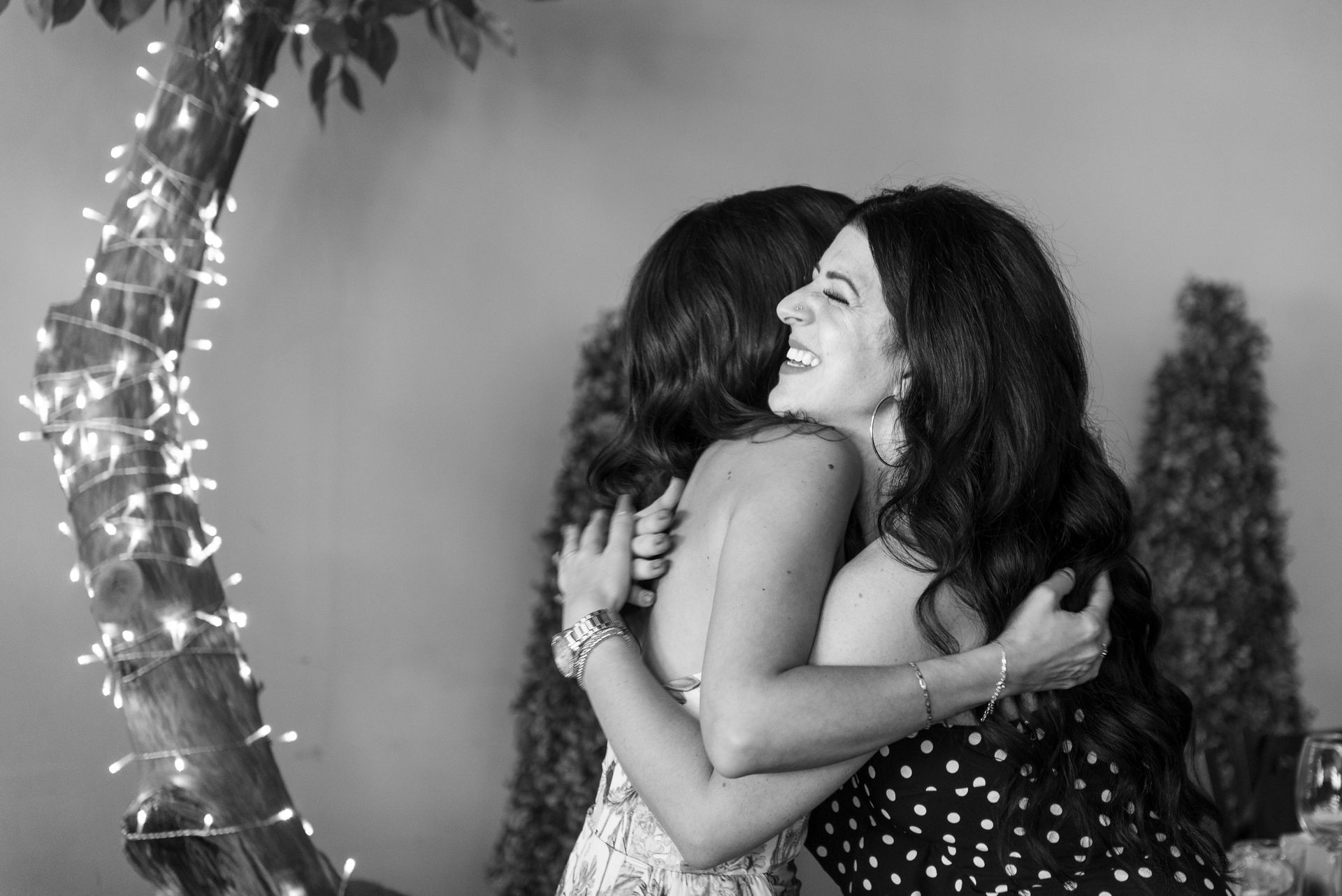 Two women embrace, smiling. One in polka dots, the other in a floral pattern, with string lights in the background.