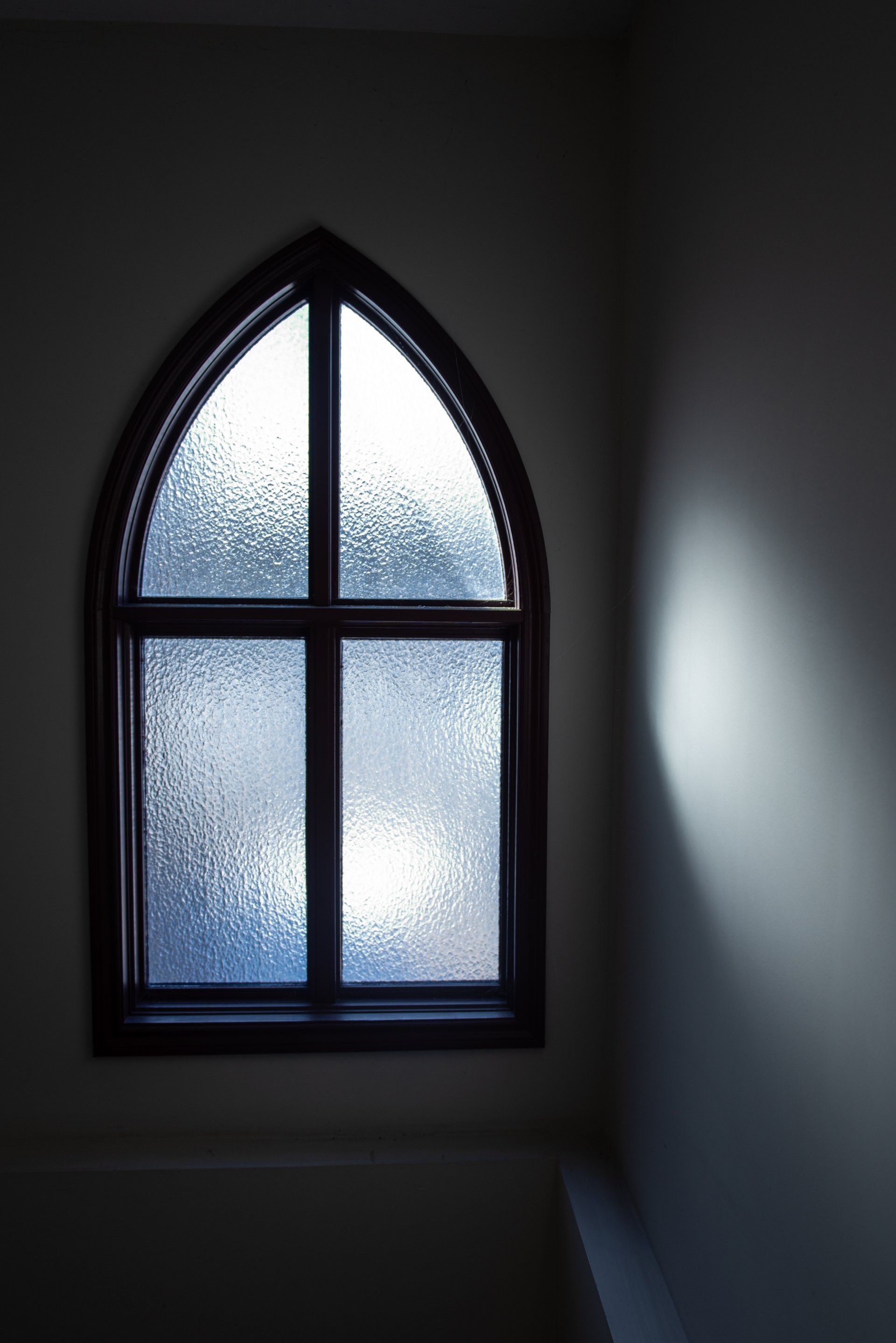 Gothic-arched window with textured glass, dark frame. Light shines from the outside, creating a bright spot on the wall.