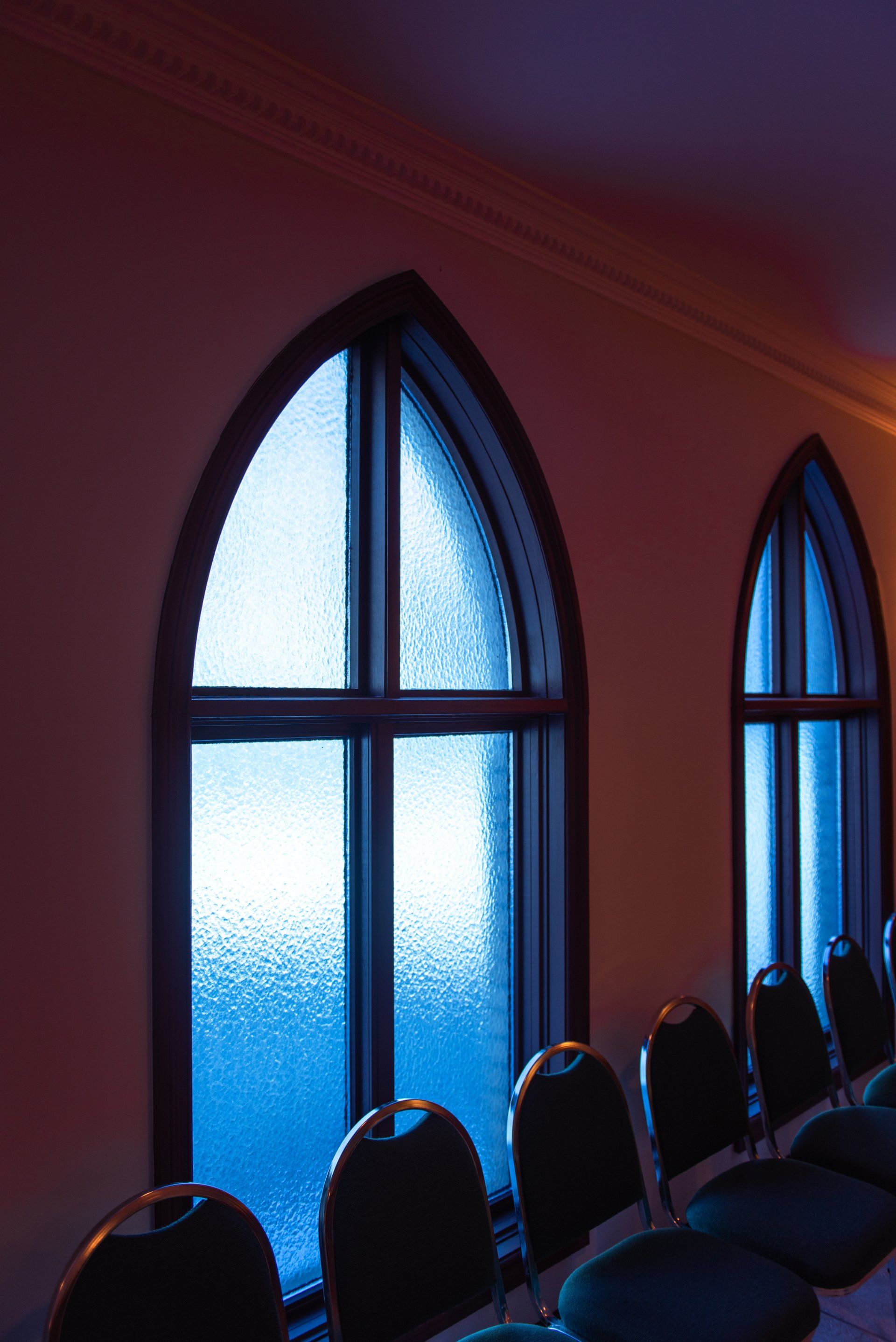 Gothic-arched windows with blue light illuminate a row of chairs in a room with warm lighting.