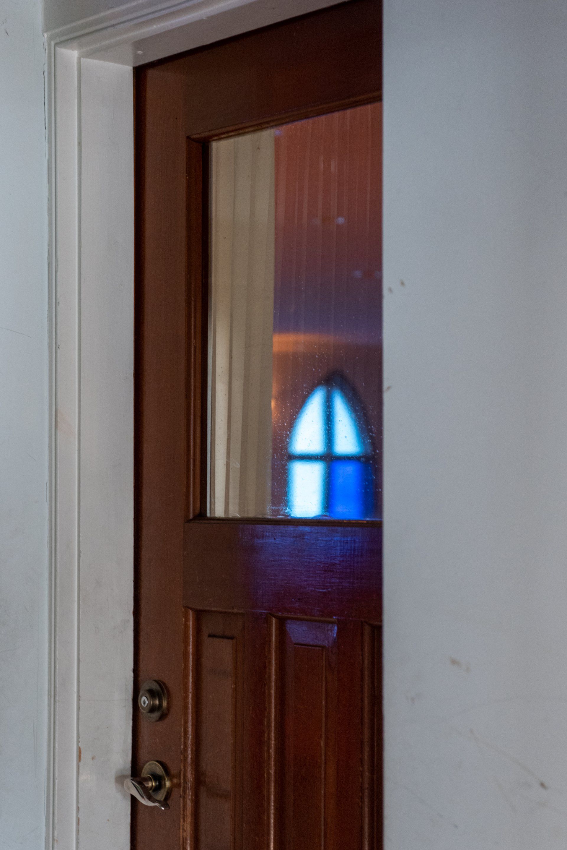 Wooden door with glass panel reflecting a blue arched window; doorframe with white walls.