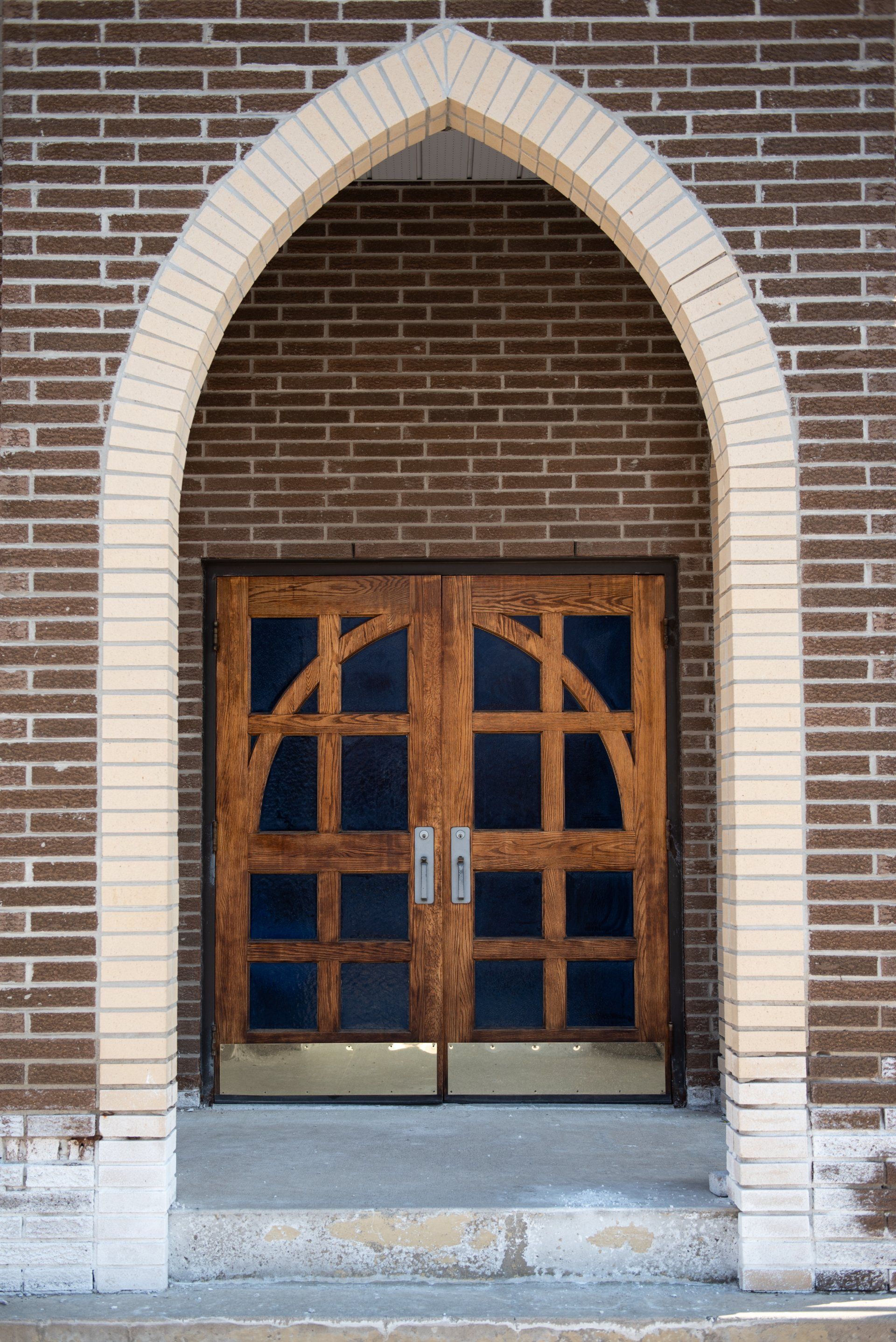 Arched doorway with wooden double doors, framed by light-colored bricks, set in a brown brick building.