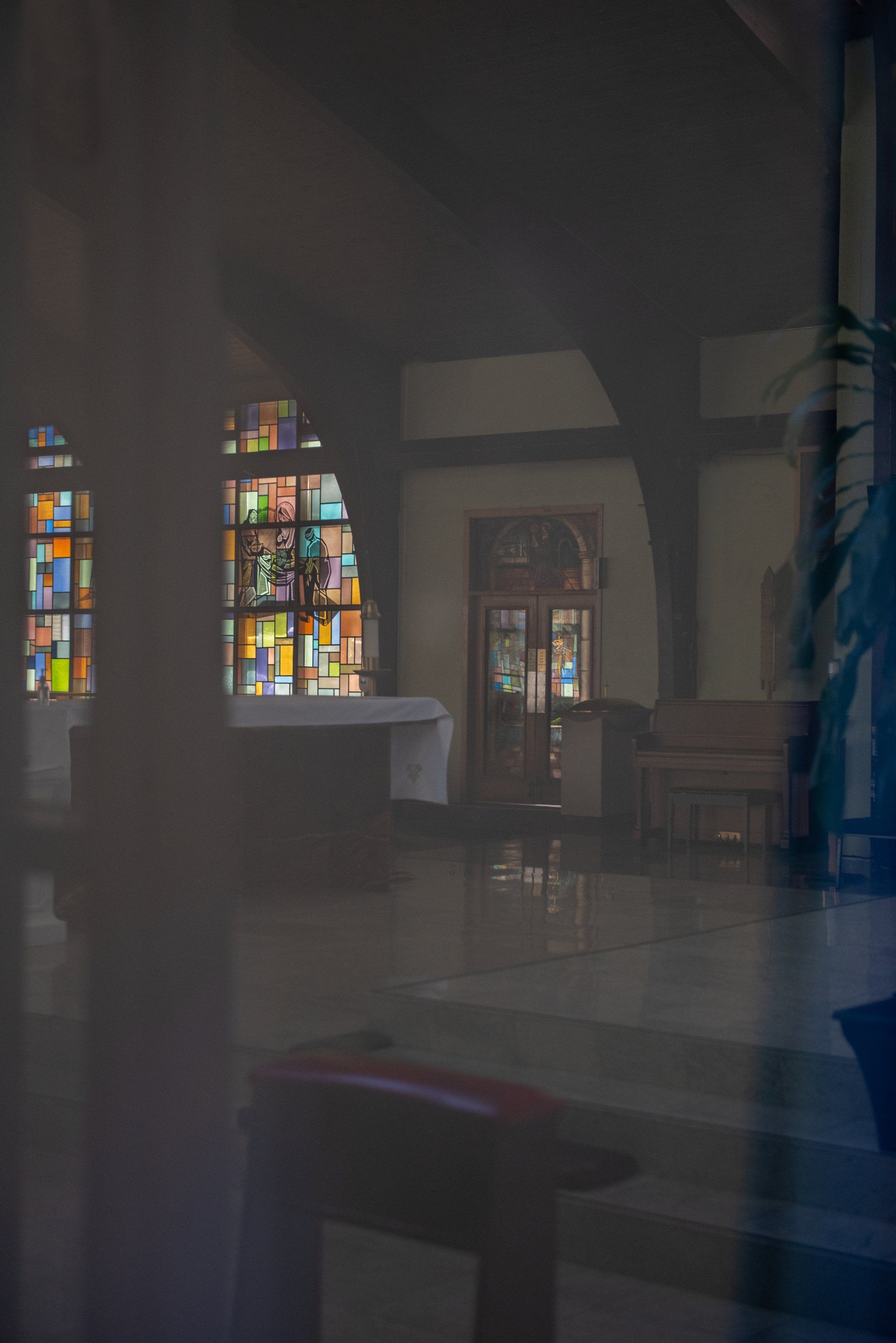 Interior church view: Stained glass windows, altar with white cloth, and wooden doors reflected in a dark surface.