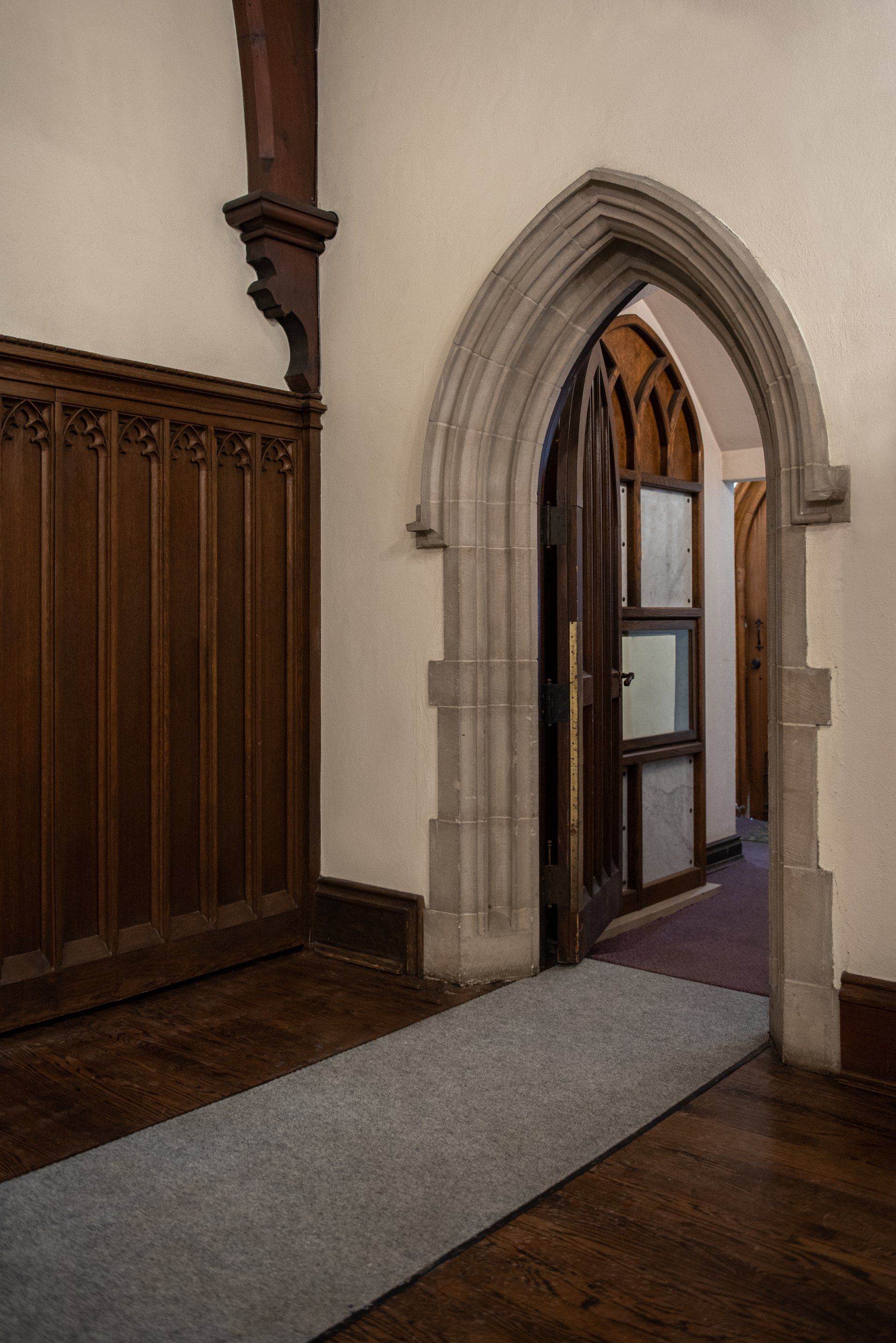 Gothic arch doorway with open door, leading to a hallway. Dark wood paneling and floor, light-colored walls. Gray runner rug.