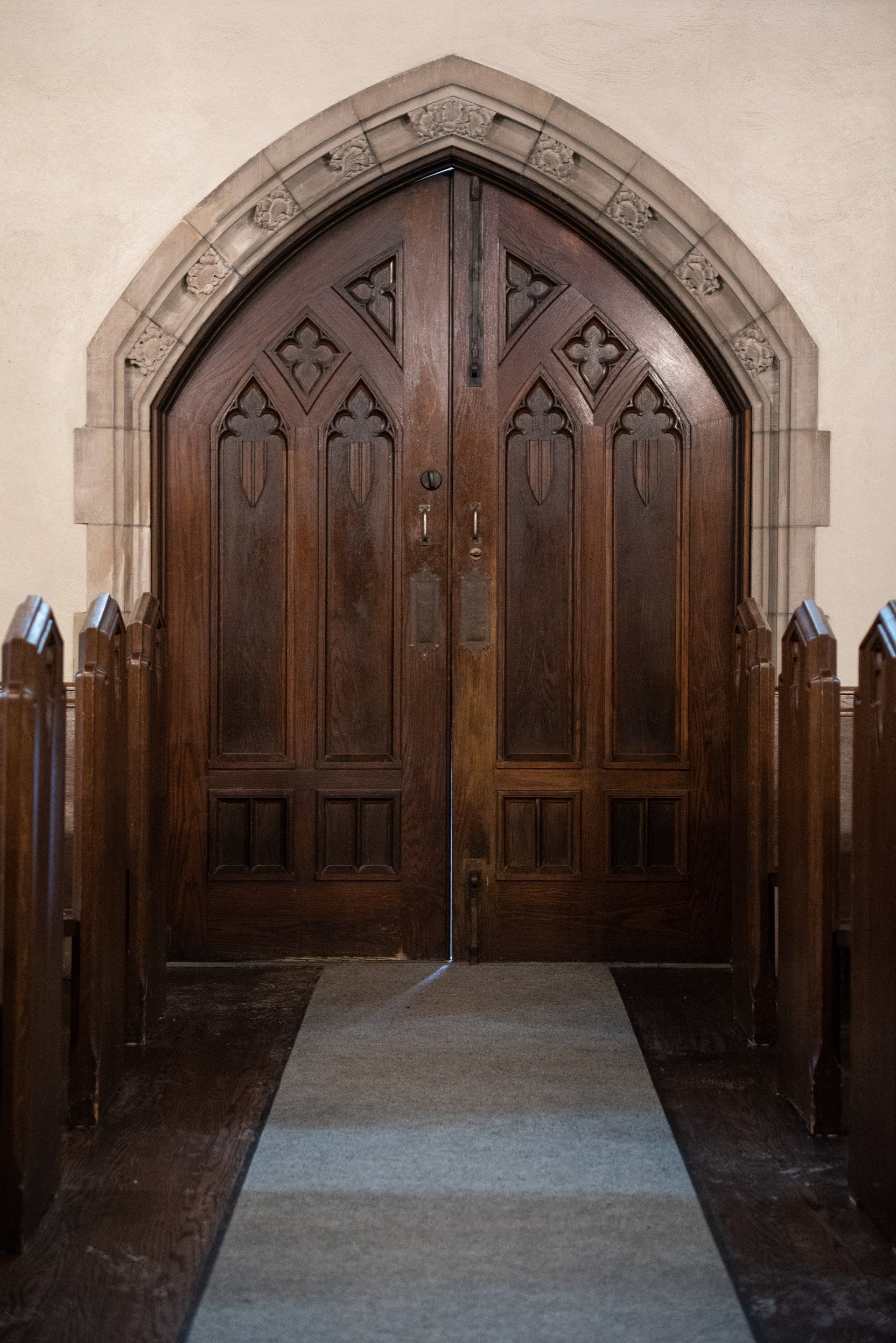 Wooden double doors with arched stone trim, leading to a church interior, flanked by pews.