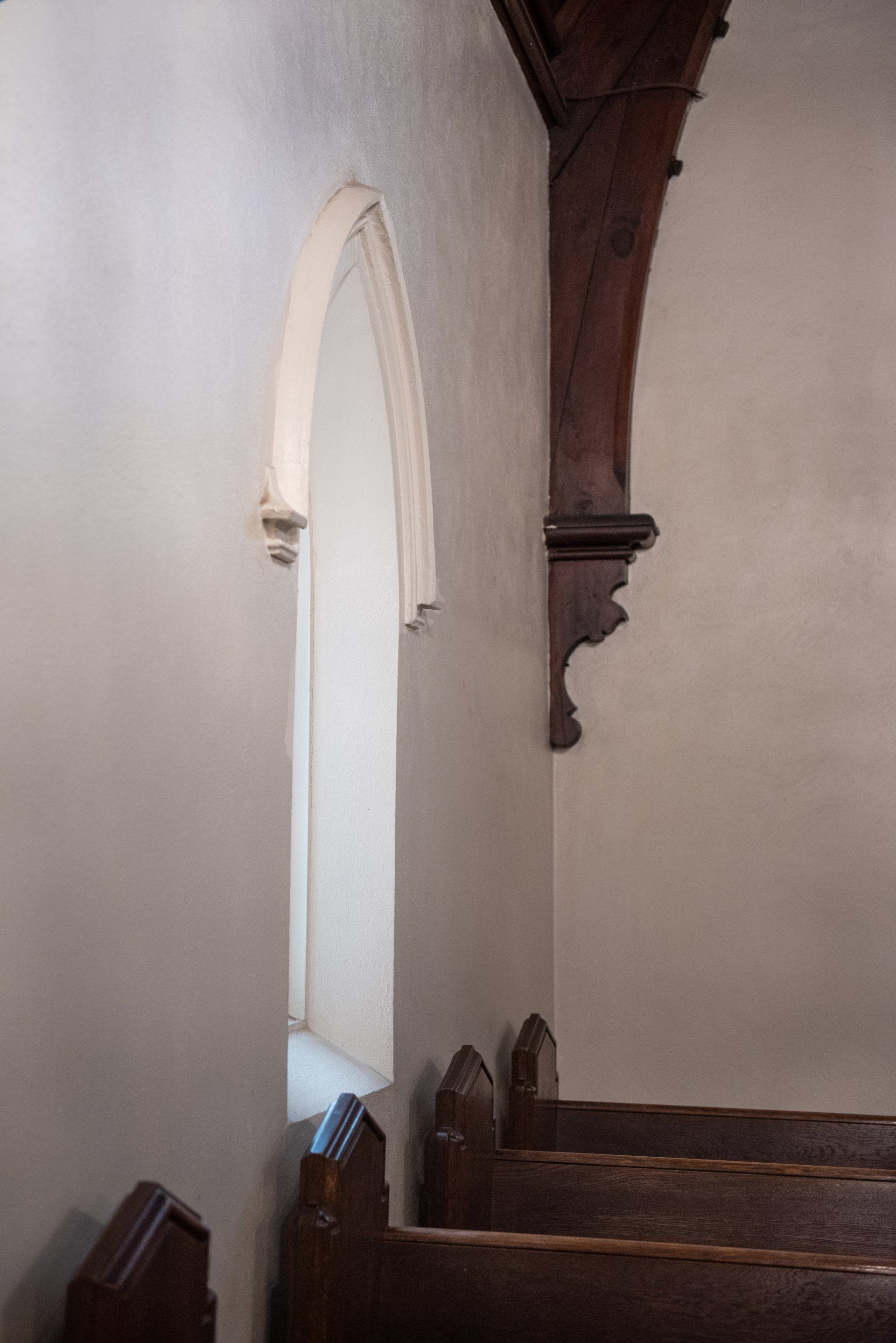 Interior corner of a church with arched window and wooden pews. Dark brown wooden support. White walls.