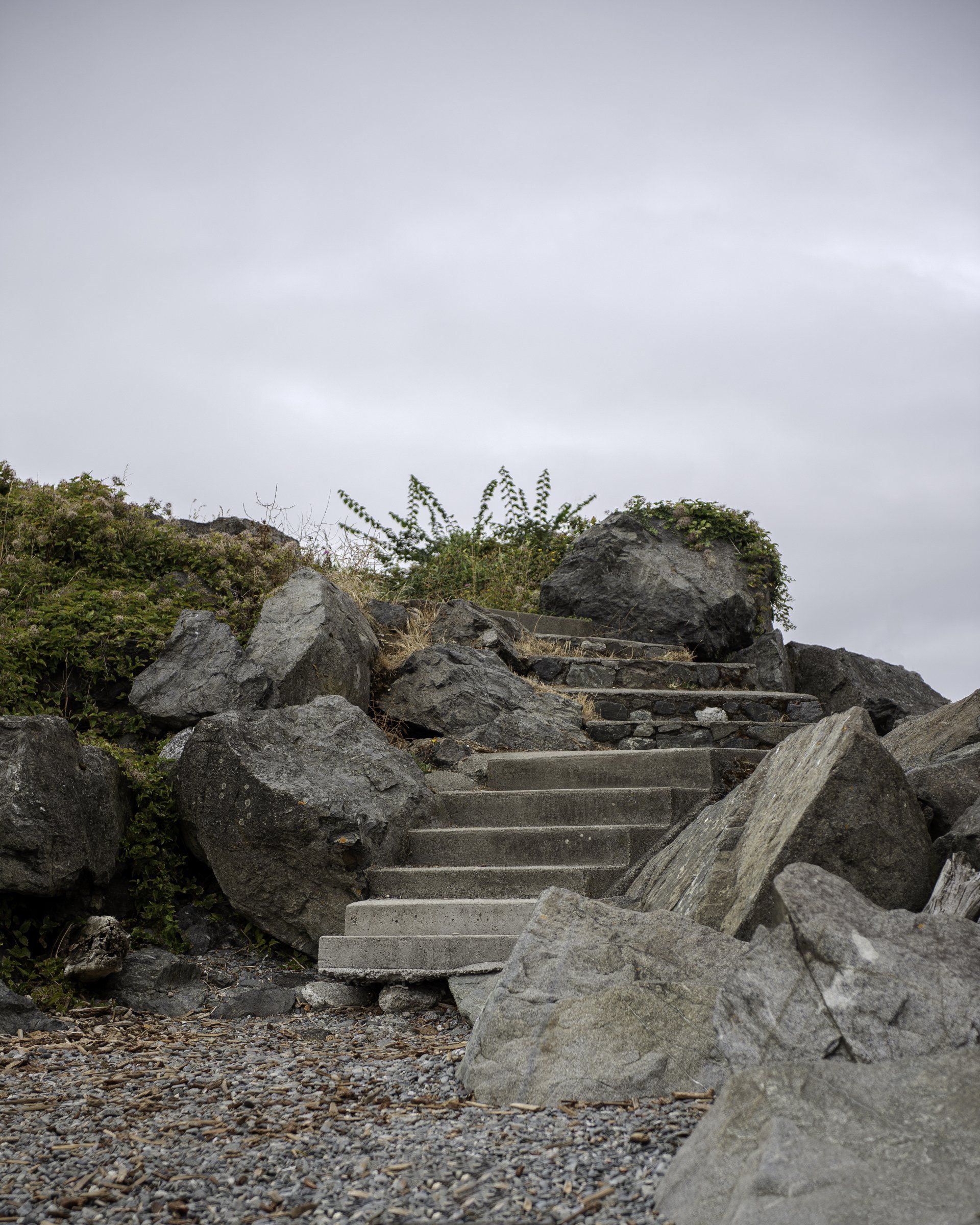 Concrete steps ascend through large rocks and foliage under a cloudy sky.
