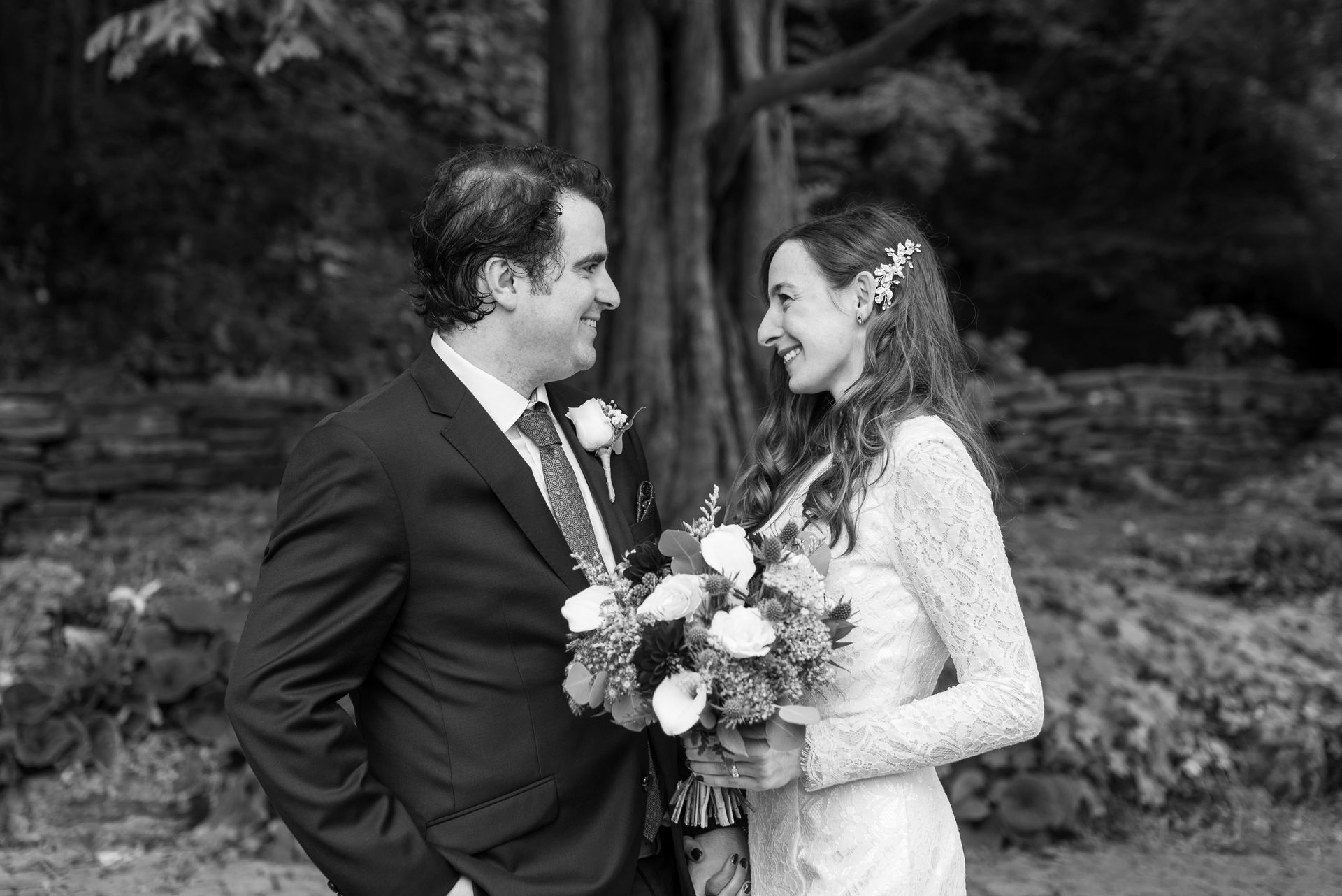 Wedding couple smiling at each other, holding flowers, standing near a tree in black and white.