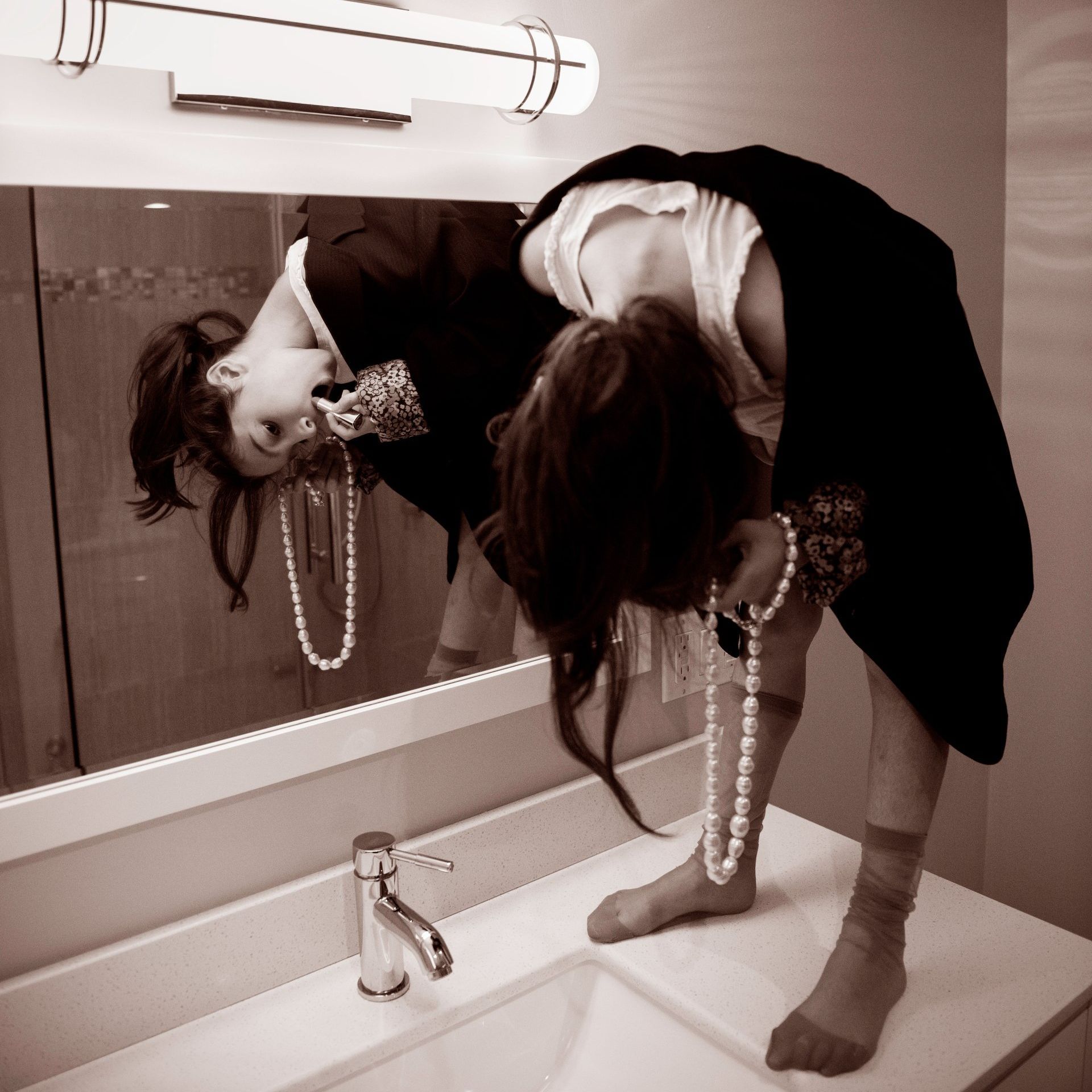 Woman bending over a bathroom sink, head reflected in mirror, holding pearl necklace.