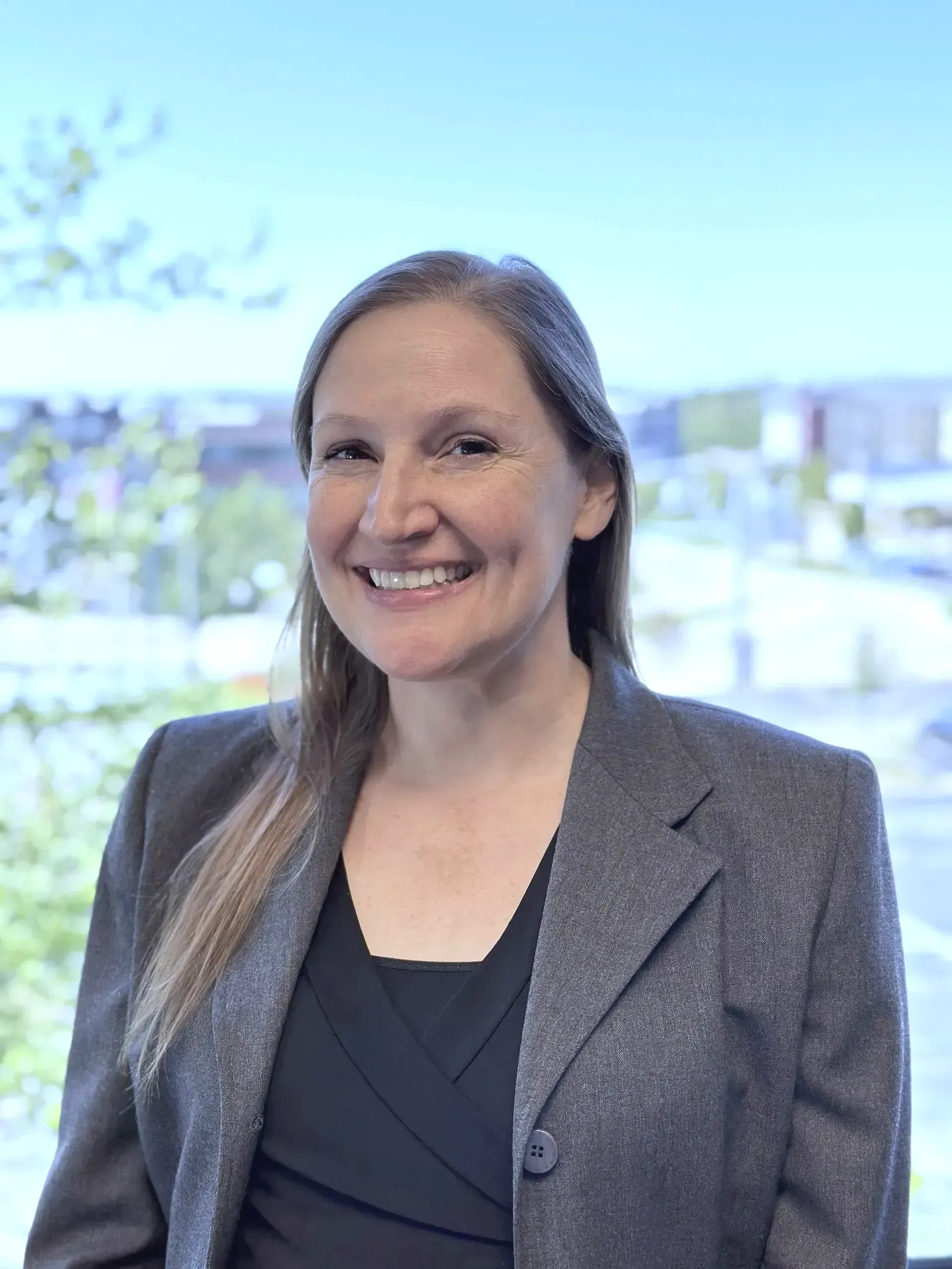 Woman with fair skin, smiling, wearing a gray blazer over a black top, in front of a window with a city view.