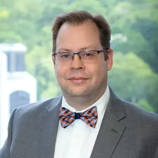 Man in glasses, bow tie, and suit jacket smiling in an office setting.