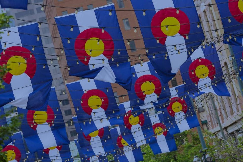 Many Colorado state flags hang suspended from string lights over an outdoor city street.