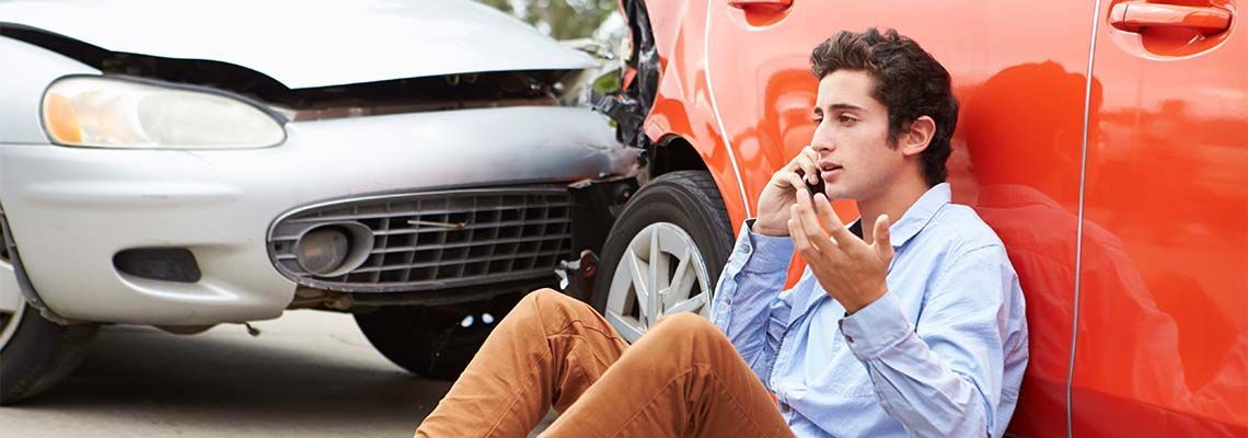 A person sits on the ground against a red car after a collision with a silver car, speaking on a phone.