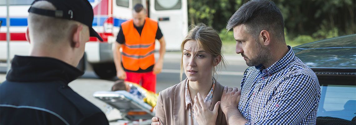 A couple looks concerned near an ambulance, while emergency responders attend to someone on a stretcher in the background.