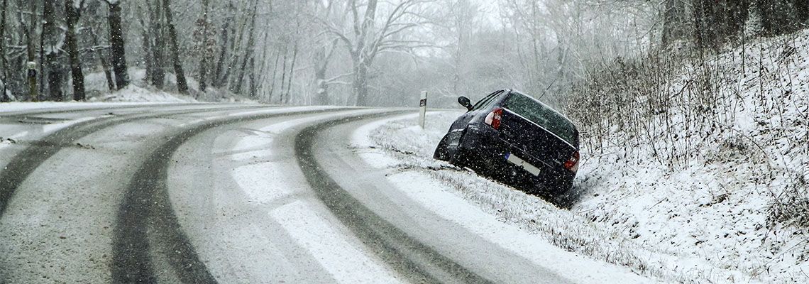 A dark-colored car stuck in the snowy roadside embankment on a curved, icy road through a wintry forest.
