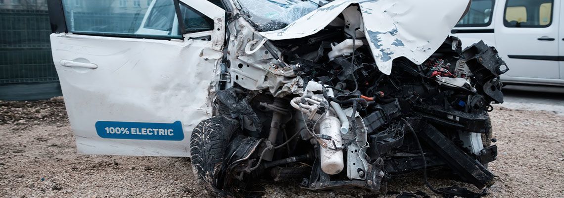 A heavily damaged white electric van sitting in a gravel lot, its front end completely crushed from a collision.