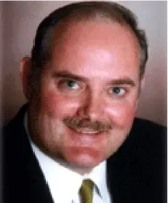 A headshot of a person with a mustache, wearing a dark suit, white shirt, and gold tie against a plain background.