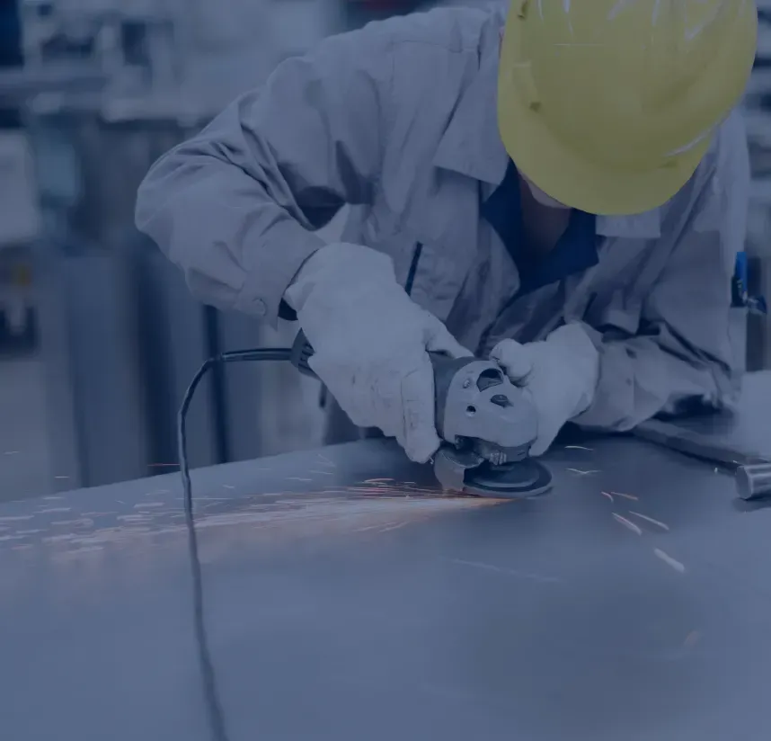 A worker in a yellow hard hat and protective gloves uses an angle grinder to cut or polish a metal surface, creating sparks.