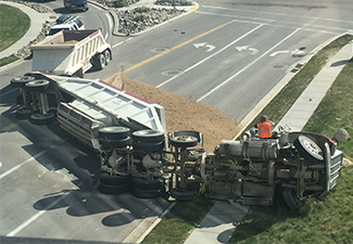 An overturned dump truck on a road intersection with its cargo of gravel spilled across the lanes.