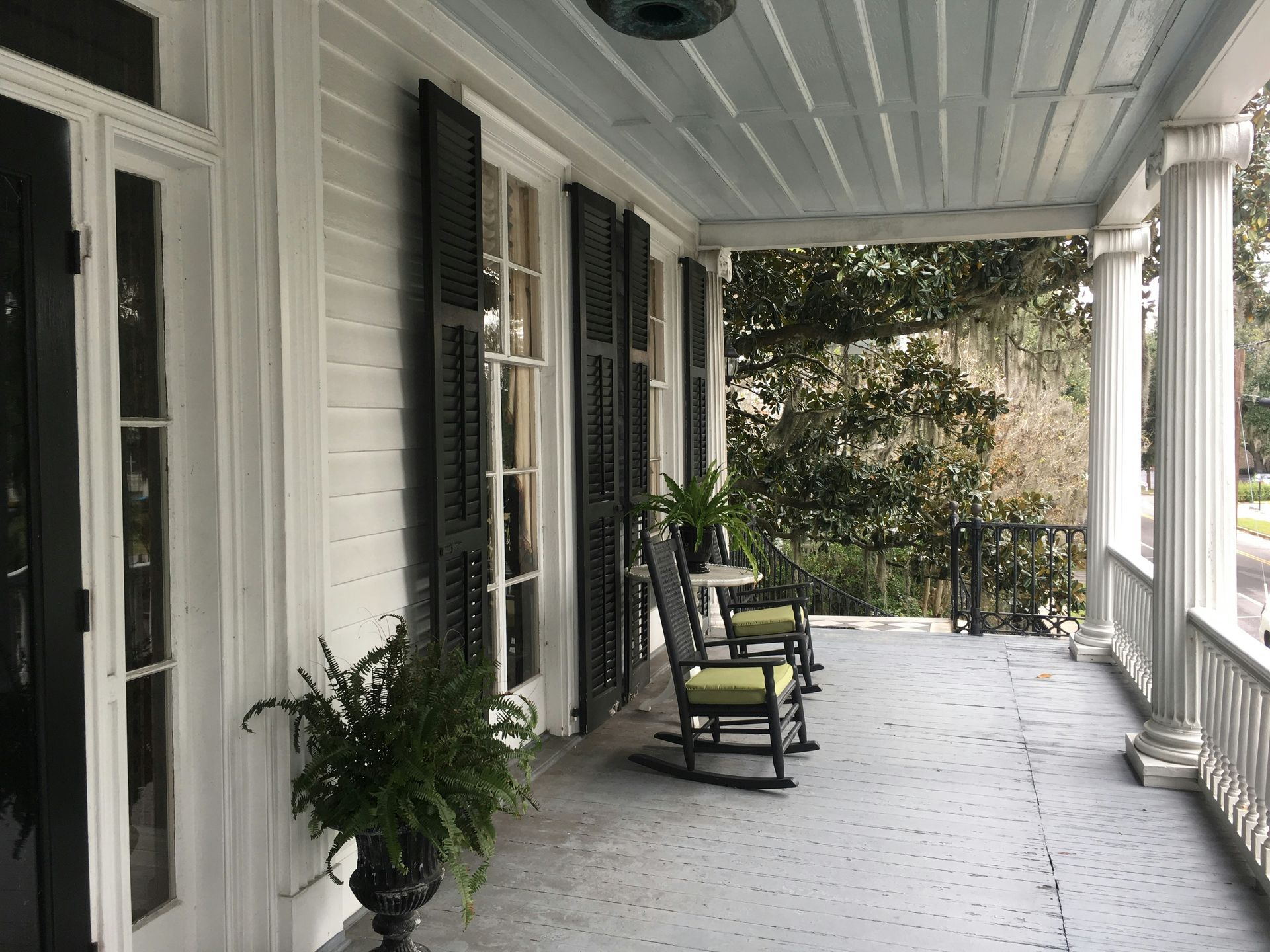 A covered front porch with white siding, black shutters, two rocking chairs, and a potted fern on a wooden floor.
