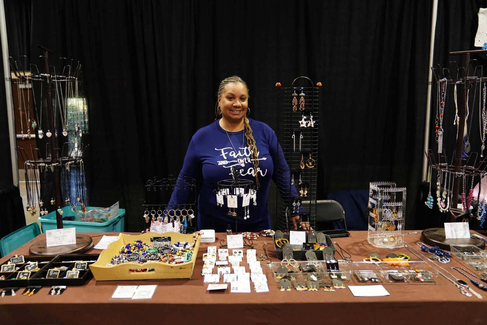 Vendor behind a table of jewelry and crafts at an indoor market booth with black drapes