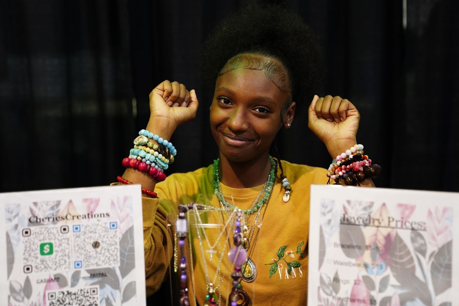 Person smiling with raised fists at a booth display, wearing colorful bracelets and a yellow shirt