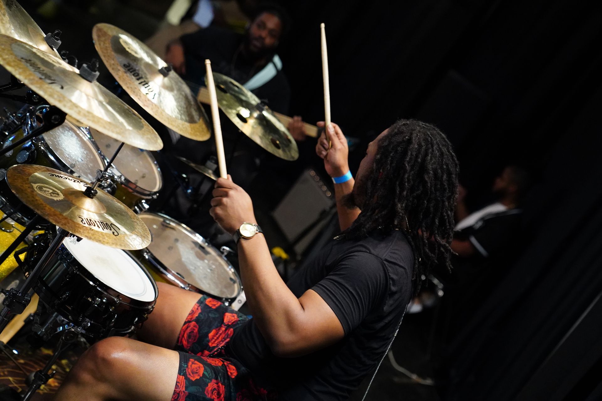 Drummer playing a drum kit onstage, with cymbals and drumsticks raised in a dark performance setting