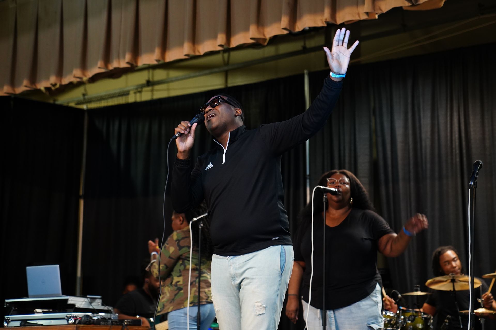 Singer on stage with raised hand, performing into a microphone beside a backup singer and drummer.
