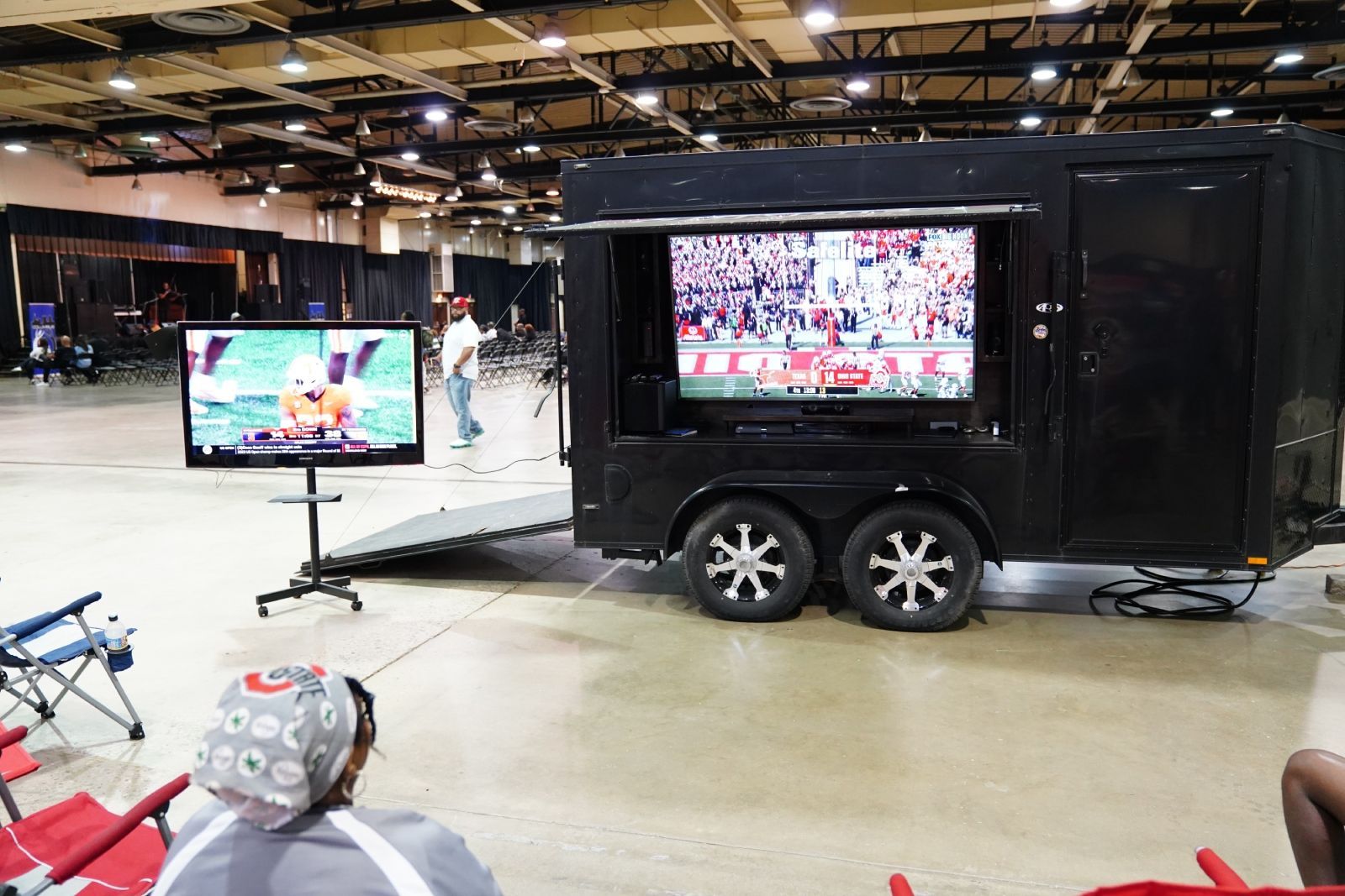 Two black trailer-mounted screens in an indoor hall, showing a live event on TVs
