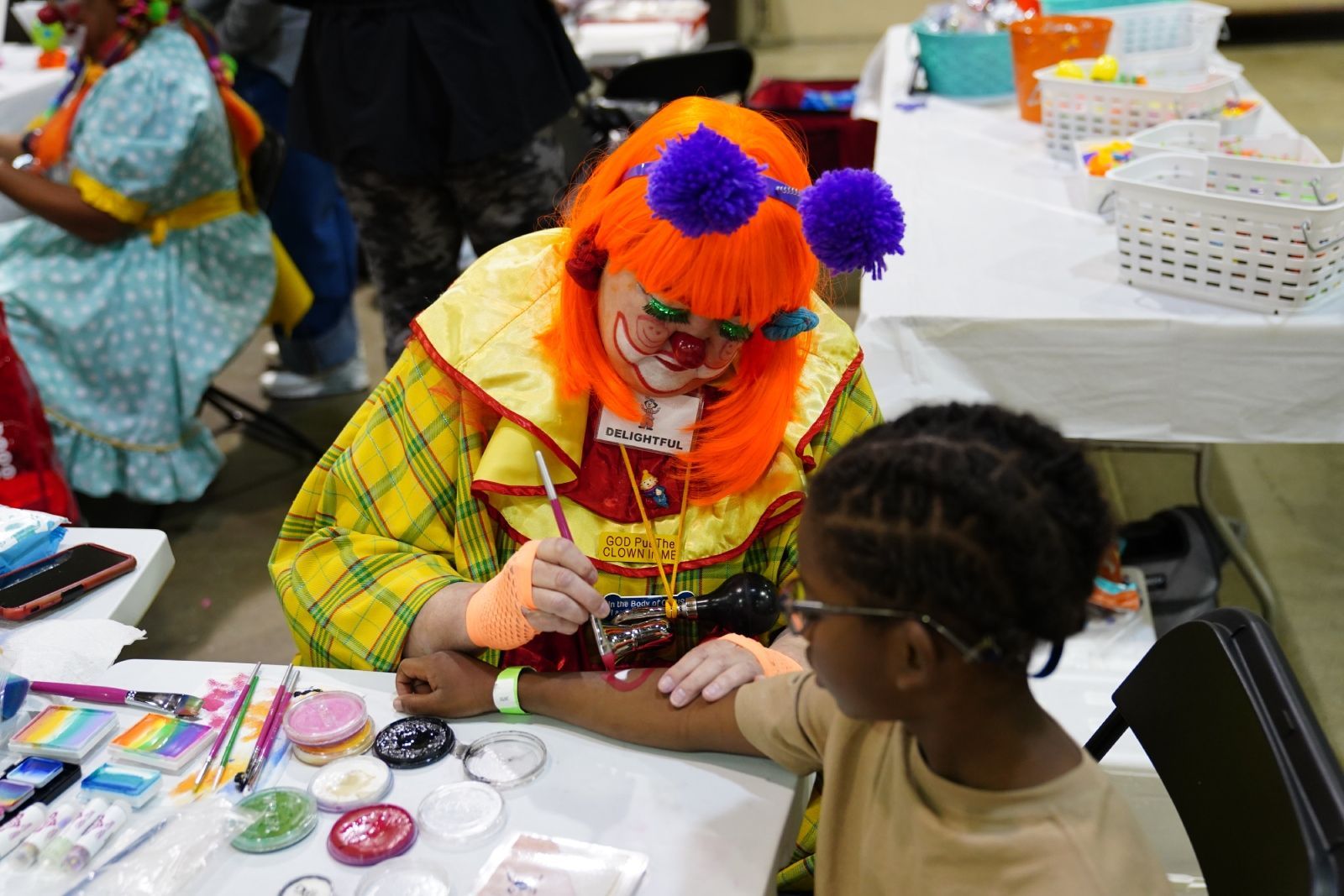 Clown painting a child’s face at a table with art supplies and a white basket nearby