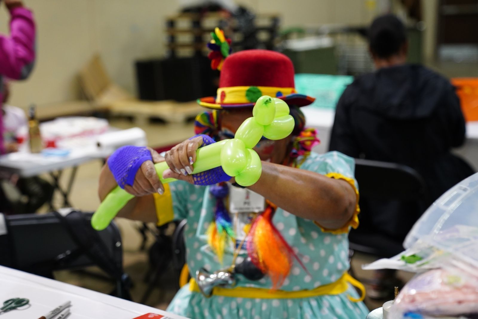 Person in red hat making a green balloon animal at a table in a busy indoor workshop
