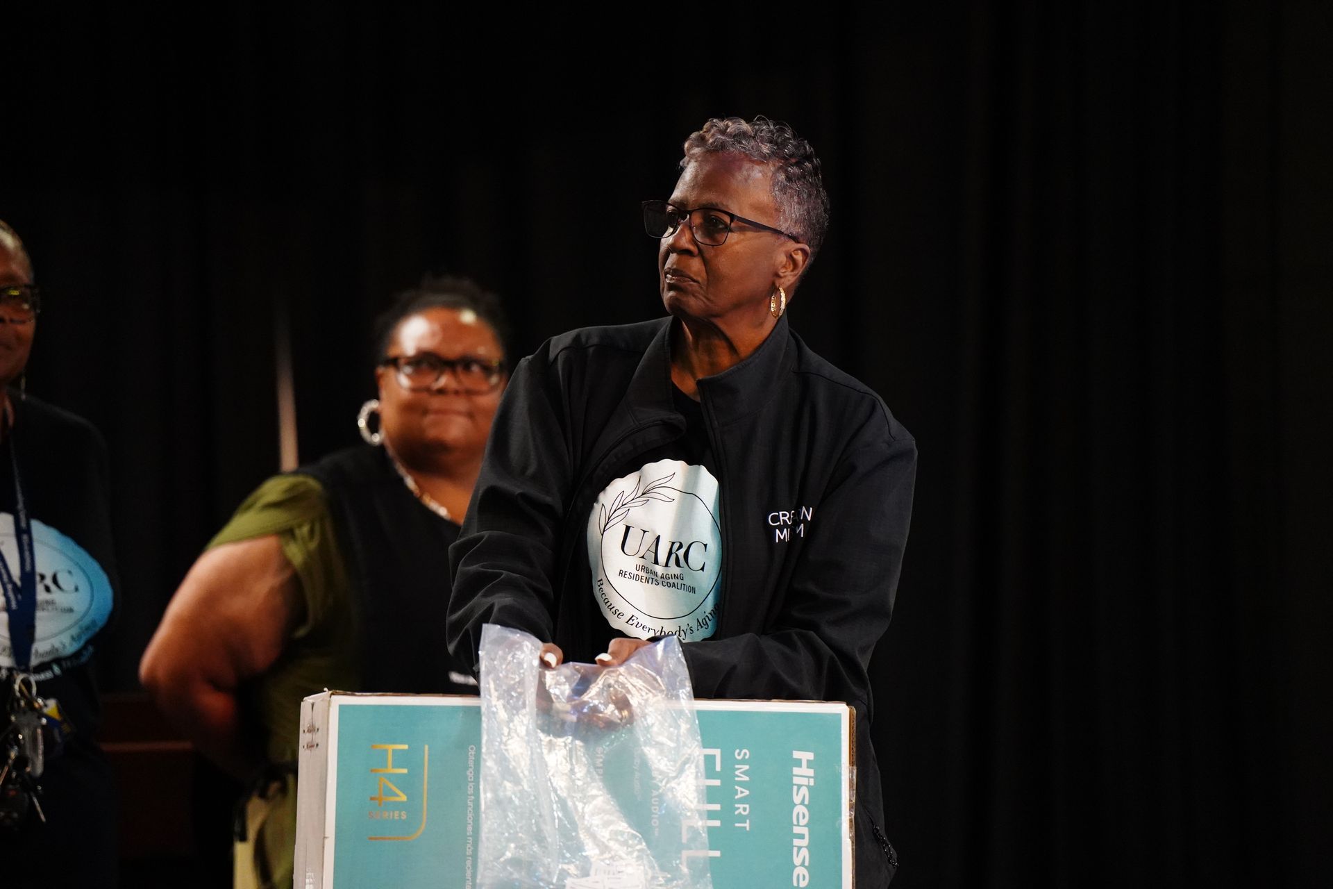 Speaker at podium on stage, wearing glasses and black shirt, with a banner draped over the front.