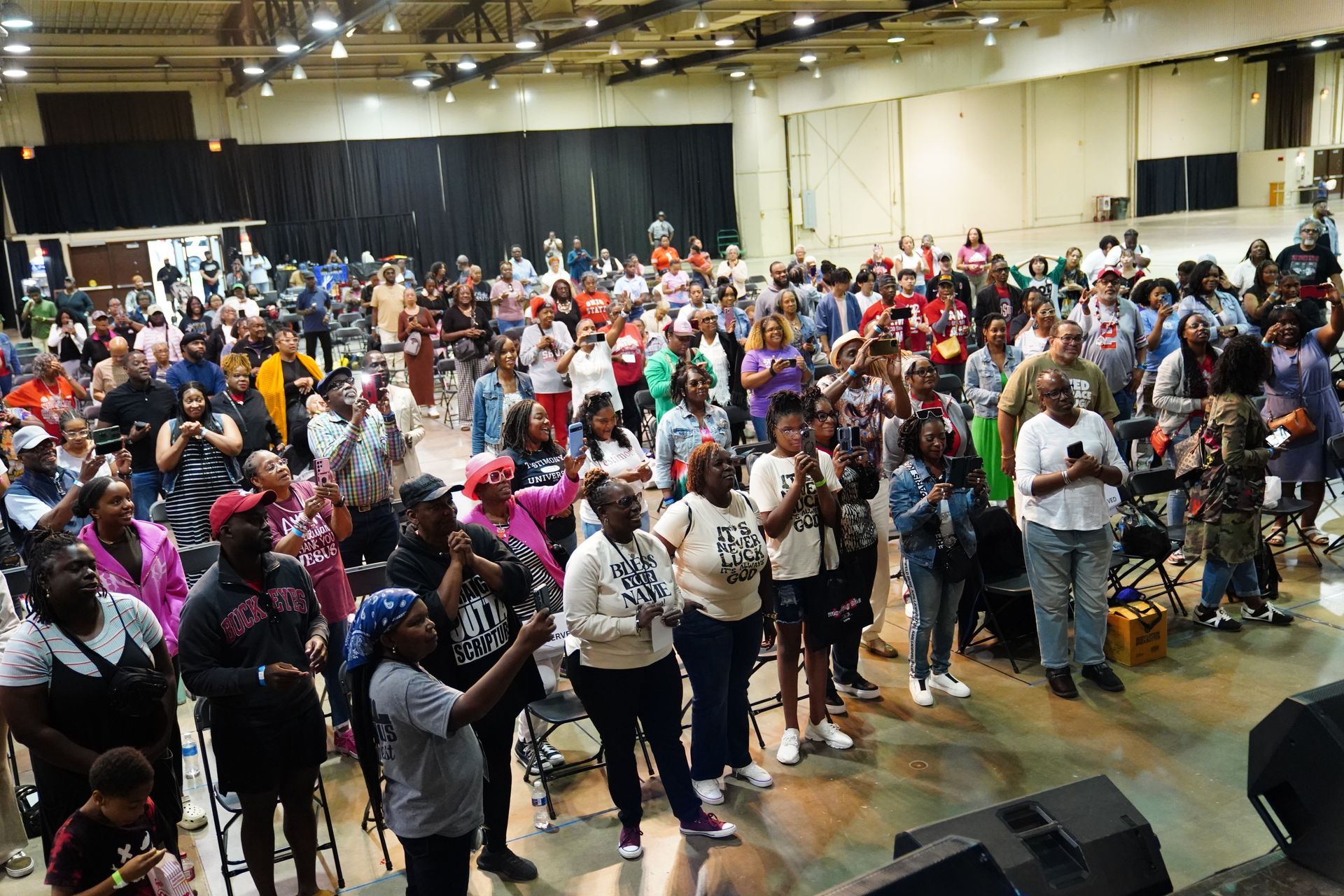 Large crowd gathered in an indoor hall, many standing and facing a stage area.