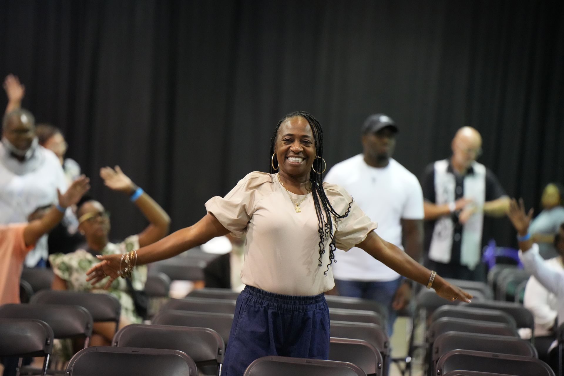 Woman smiling with arms outstretched in an auditorium, with seated people blurred in the background