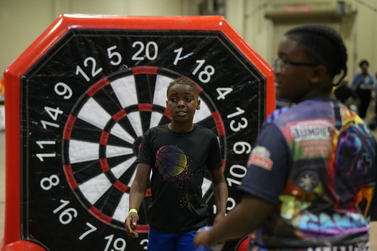 Two people in black shirts stand before a dartboard at an indoor game event.