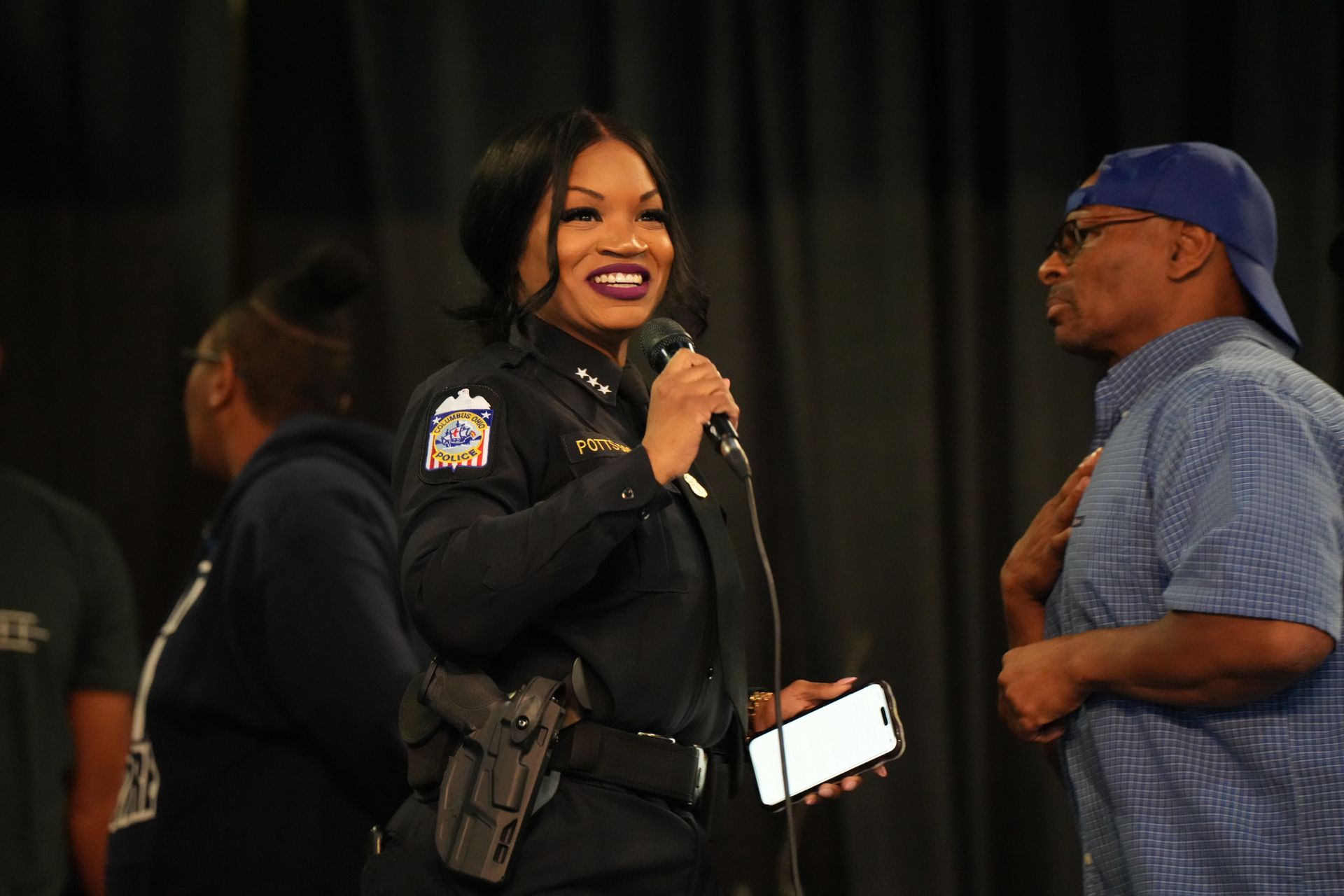 Woman in black uniform speaking into a microphone beside a man in a blue cap on a stage