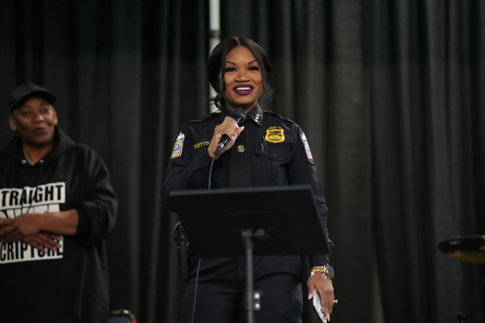 Speaker at podium in dark uniform, smiling and holding a microphone onstage before a black curtain