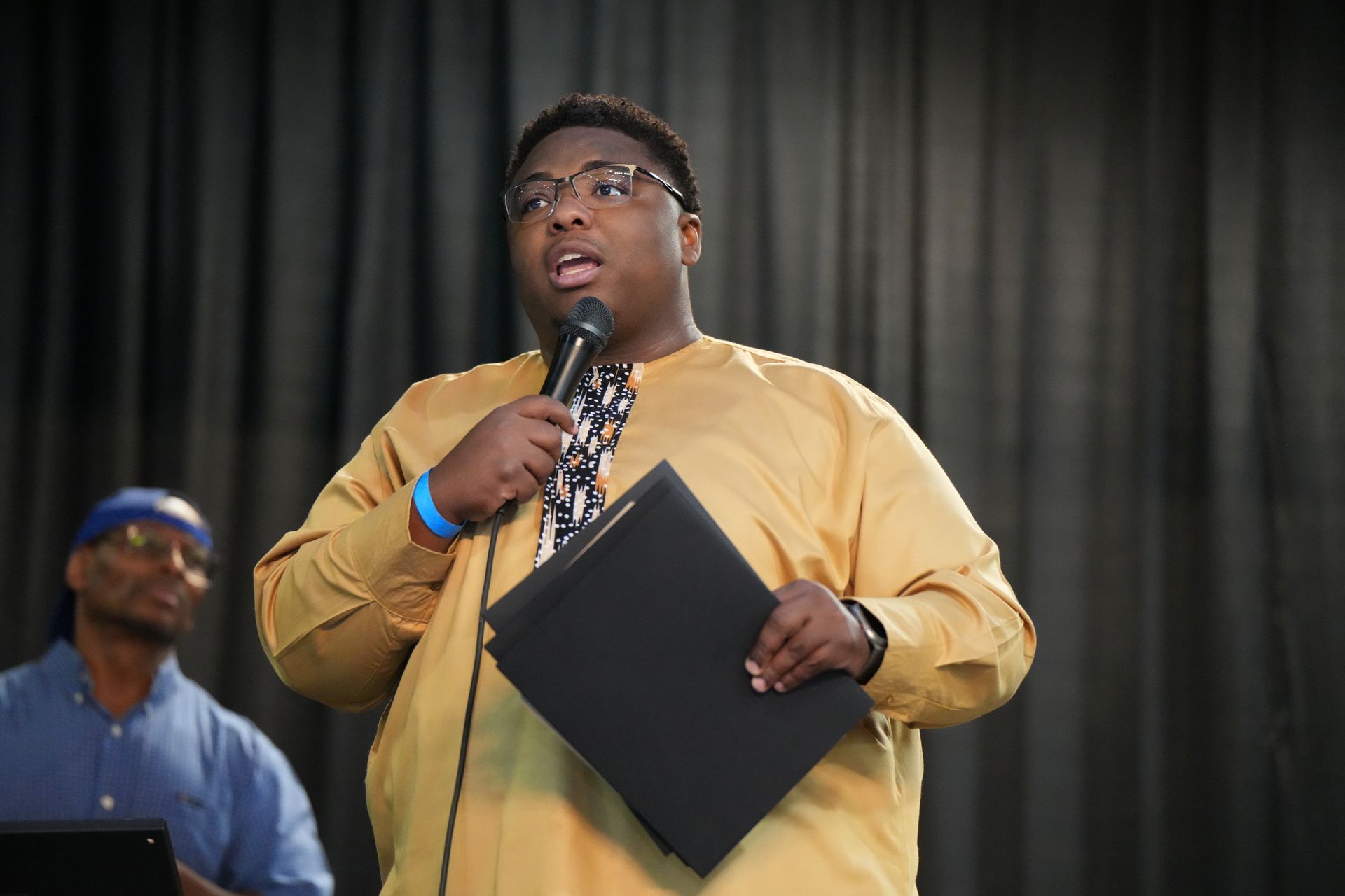 Speaker holding a microphone and black folder on stage in front of dark curtains