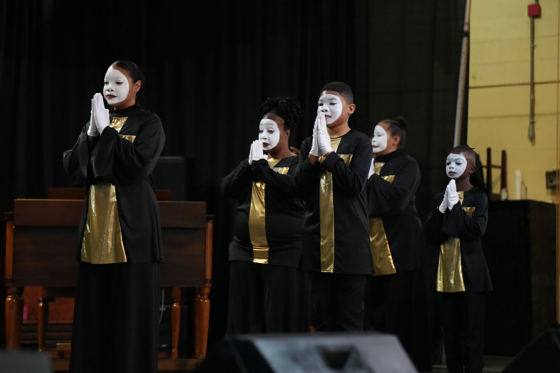 Four performers in black and gold stand on stage holding white masks to their faces.