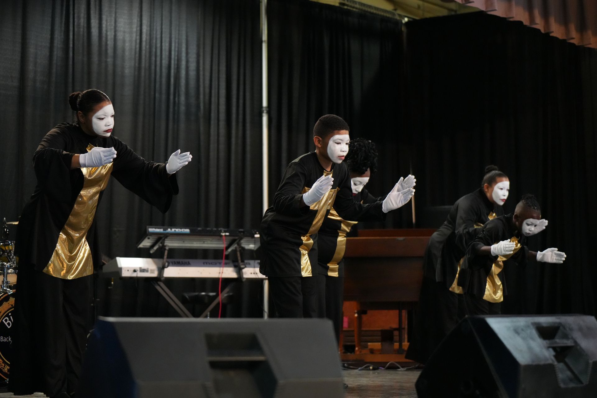 Three masked performers in black and gold costumes pose on a stage with microphones.